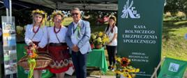 Stoisko informacyjno-prewencyjne podczas Dożynek Gminno-Parafialnych w Sułoszowej. Przed namiotem z logo KRUS znajdują się panie z KGW w strojach regionalnych, obok nich stoją pracownicy Placówki Terenowej KRU w Wolbromiu. Przed namiotem wystawiono bukiet ze słoneczników oraz leżak.