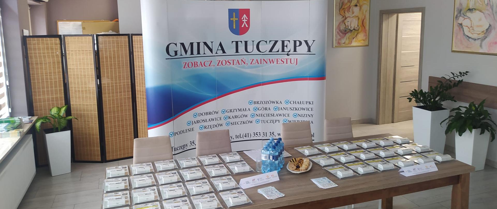 Czujki tlenku węgla leżące na stole, W tle baner gminy Tuczępy. Na ścianie wiszą obrazy. 