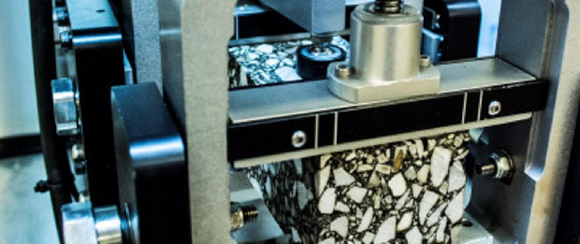 Na zdjęciu widoczna jest maszyna laboratoryjna używana do testowania wytrzymałości materiałów. W centralnej części urządzenia znajduje się próbka materiału o nieregularnej strukturze, przypominającej mozaikę, która jest poddawana naciskowi przez metalowy tłok. Maszyna składa się z solidnych metalowych ram i precyzyjnych mechanizmów, które umożliwiają dokładne pomiary siły działającej na próbkę. Całość znajduje się w sterylnym środowisku laboratoryjnym.