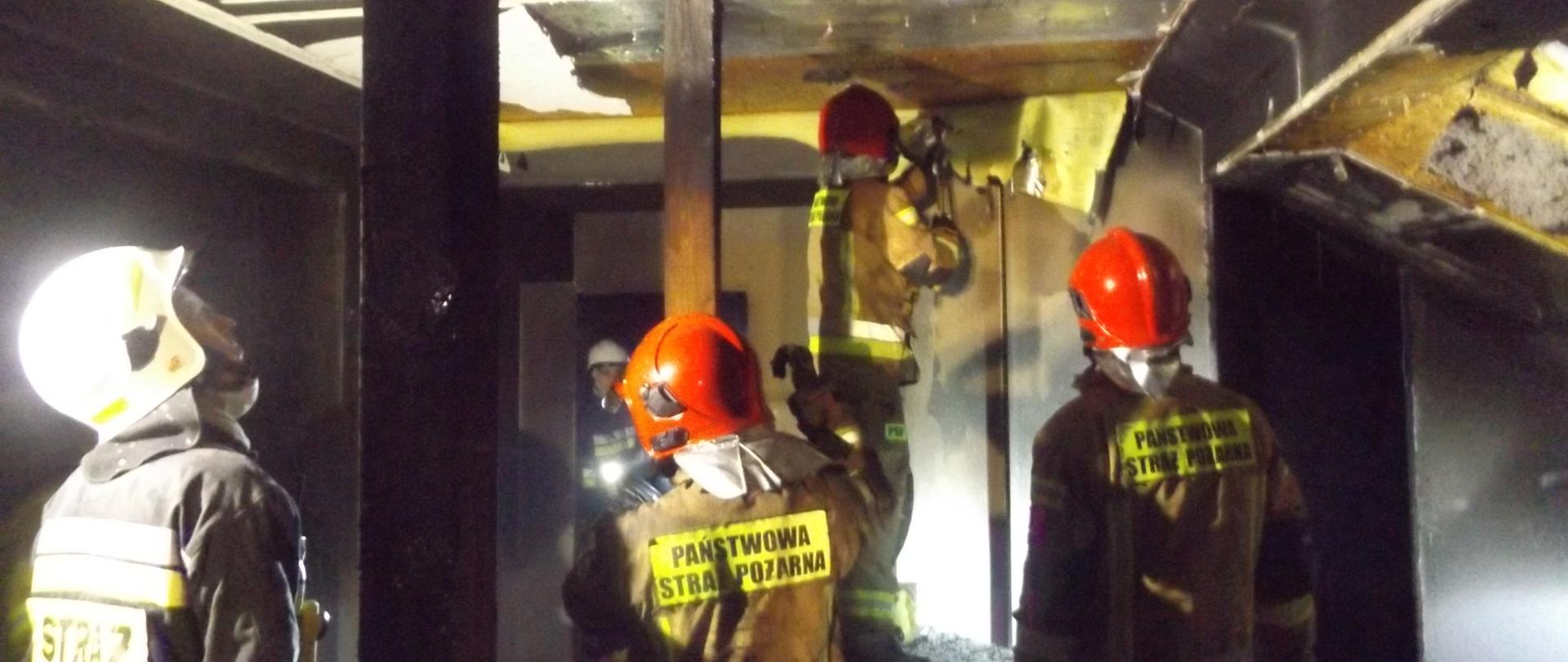 Na zdjęciu widać pięciu strażaków, którzy pracują wewnątrz pomieszczenia w którym wybuchł pożar. Strażacy przy użyciu sprzętu burzącego dokonują rozbiórki spalonych elementów wykończeniowych poddasza.