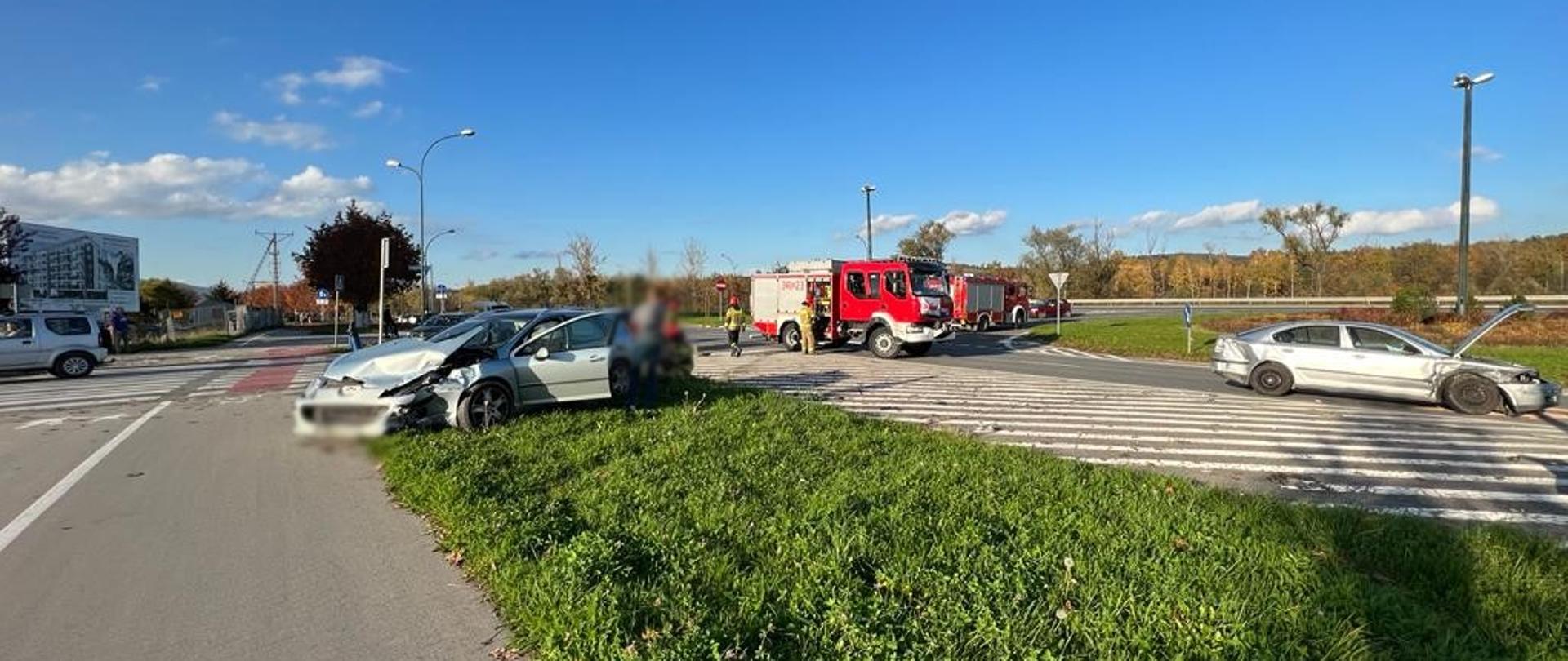 Jeden samochód osobowy stoi na pasie zieleni a drugi na drodze. W tle samochód pożarniczy.