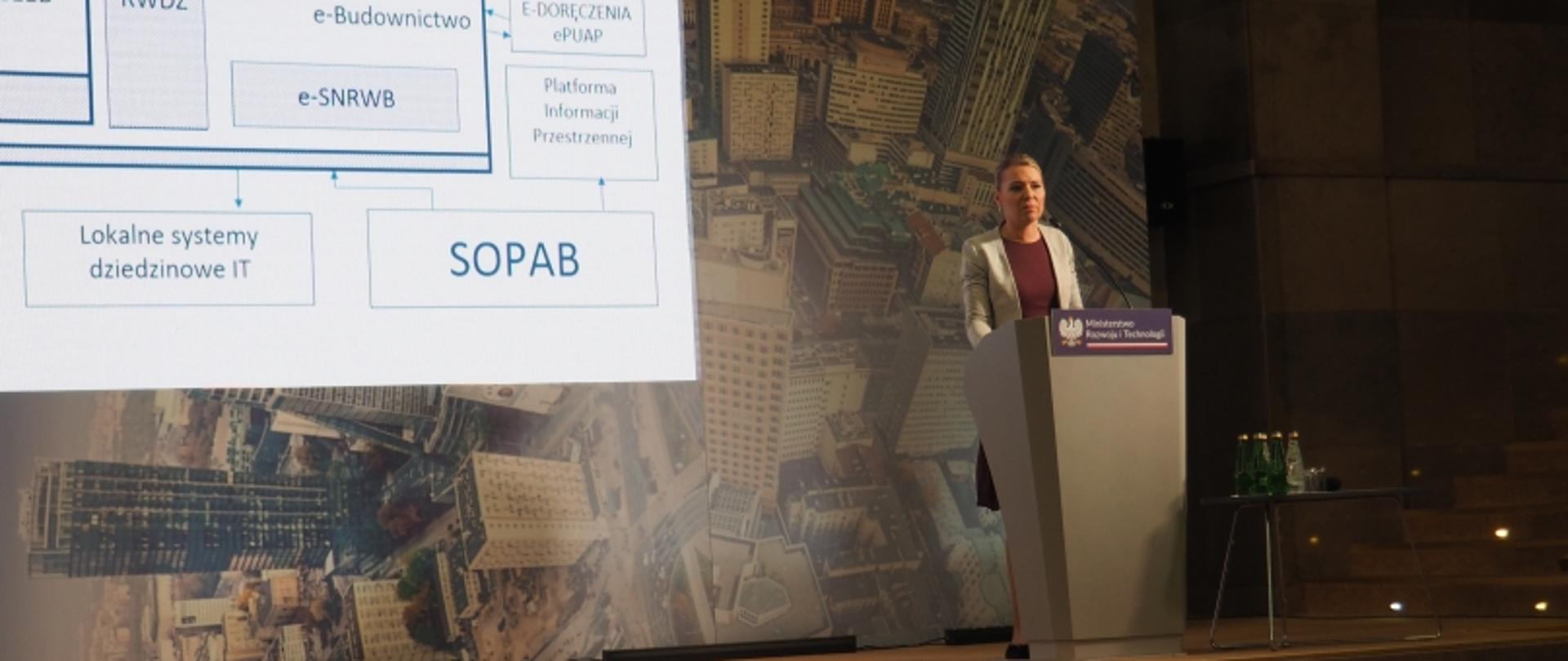 Osoba prezentująca przy mównicy Ministerstwa Rozwoju i Technologii; w tle ekran z diagramem systemów IT, takich jak e-KWZD, e-Budownictwo, e-PUAP i inne.