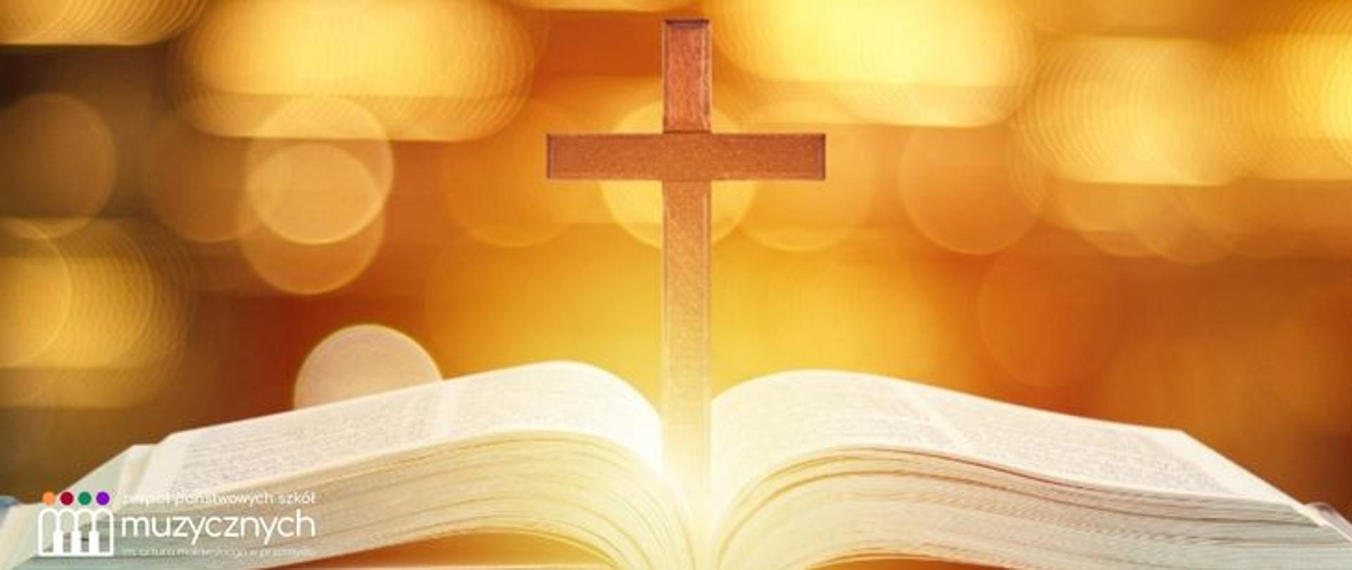 Zdjęcie przedstawiające otwartą Biblię, nad którą stoi drewniany krzyż. Tło jest ciepłe, złote i rozmyte, z efektem świetlnych refleksów, tworzących nastrojową, sakralną atmosferę. W lewym dolnym rogu widoczne logo zespołu państwowych szkół muzycznych.
