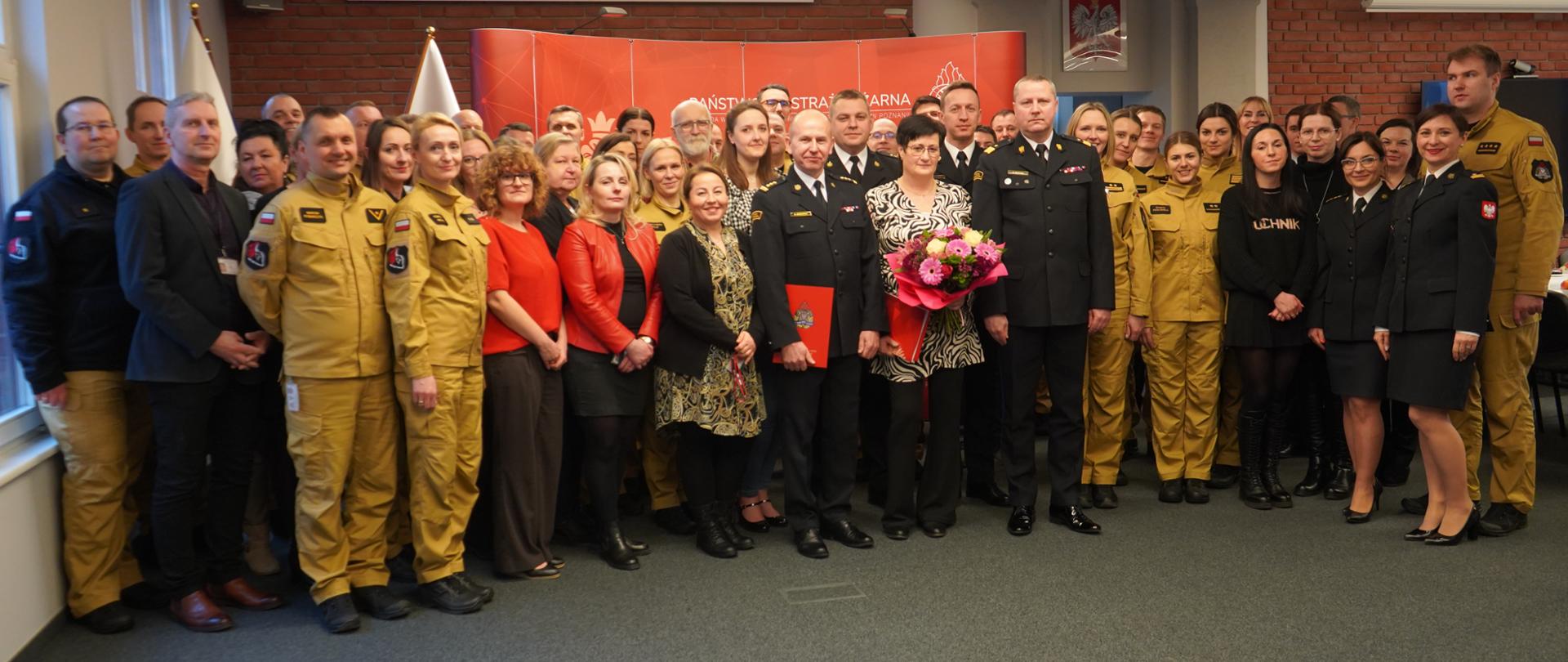 zdjęcie grupowe funkcjonariuszy i pracowników cywilnych , w tle ściana z cegły i flagi biało czerwone 