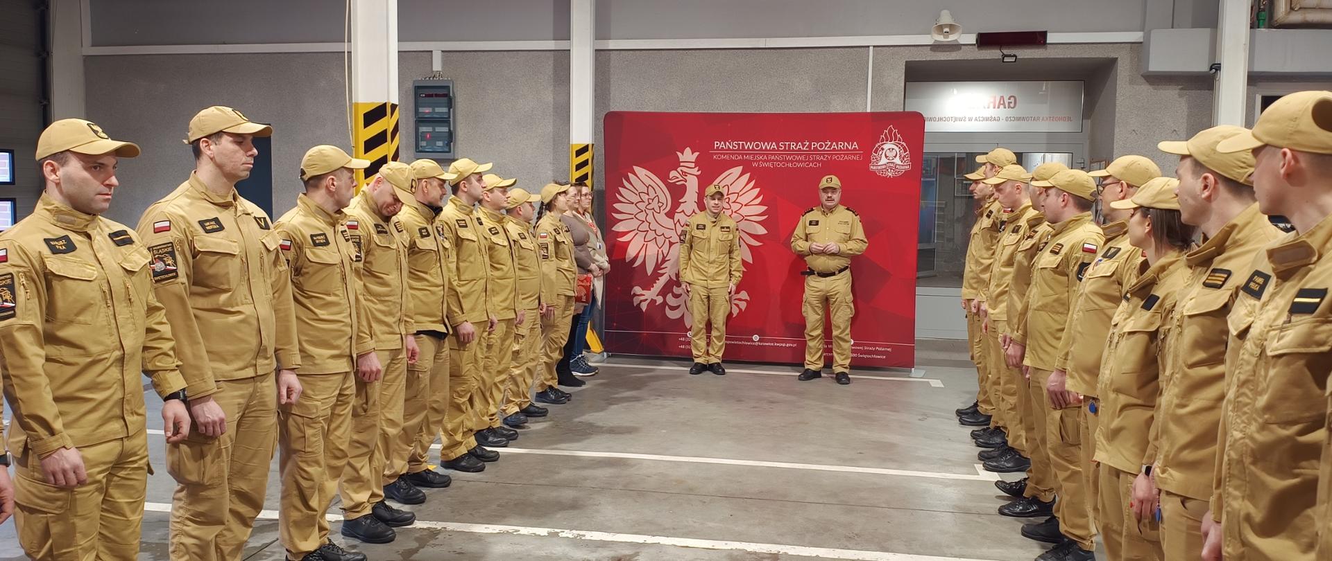 Zdjęcie przedstawia zmianę służby funkcjonariuszy Państwowej Straży Pożarnej wewnątrz garażu. Strażacy, ustawieni w dwóch równoległych szeregach, mają na sobie piaskowe mundury służbowe oraz czapki z daszkiem w tym samym kolorze. W tle, pomiędzy formacjami, widoczna jest czerwona ścianka ekspozycyjna z godłem Polski (orłem w koronie) oraz logo straży pożarnej, przy której stoi dwóch dowódców prowadzących odprawę lub uroczystość.