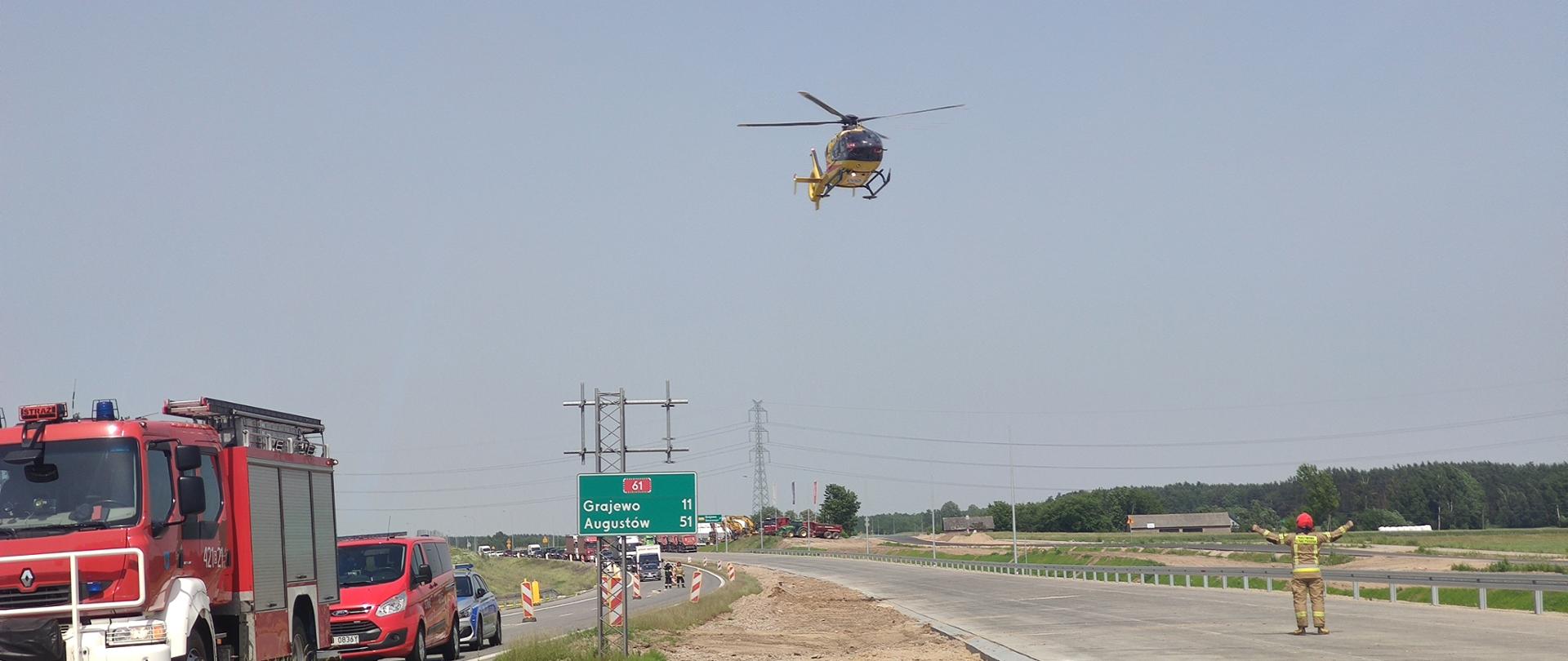 Śmigłowiec LPR w trakcie lądowania na jezdni. Z prawej strony stoi strażak i sprowadza gestem lądowania śmigłowiec. Z prawej stoją pojazdy służb ratowniczych.