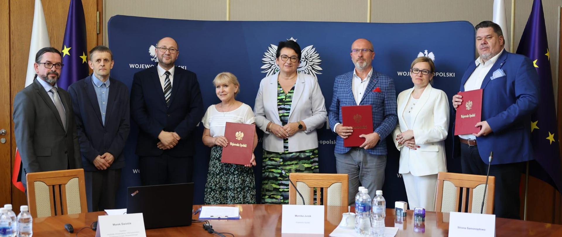 Wojewoda Opolski Monika Jurek razem z wójtami gmin: Walce, Skoroszyce, Reńska Wieś i Głuchołazy, po podpisaniu umów o dofinansowanie ze środków z Rządowego Funduszu Rozwoju Dróg. Na zdjęciu Wojewoda Opolski stoi na środku. Wszyscy stoją przed stołem, na którym leży między innymi laptop, butelki wody, dokumenty czy pieczątki. W tle widać również flagi Polski oraz Unii Europejskiej. 