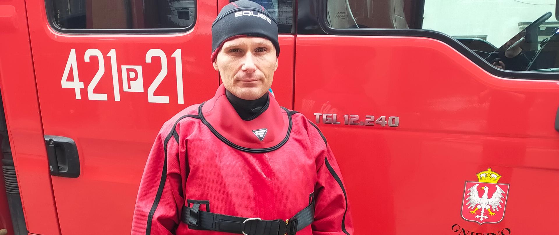 Na zdjęciu widać strażaka w ubraniu wodno-lodowym koloru czerwonego. Stoi on przy samochodzie strażackim