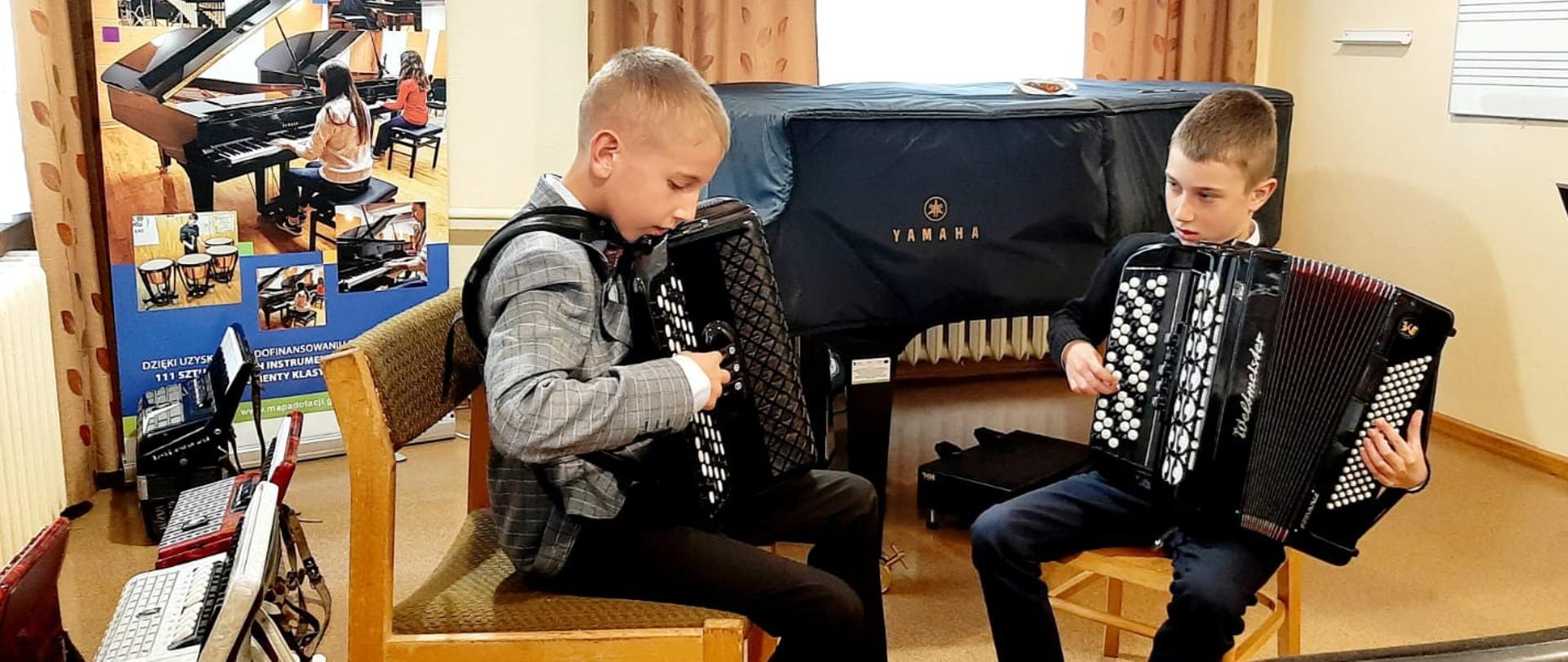 Uczniowie podczas audycji akordeonowej grają na dwóch akordeonach siedząc na przeciw siebie. Za nimi fortepian przykryty pokrowcem oraz okno przysłonięte zasłonami. Z lewej strony na podłodze leżą 4 różne akordeony a za mi baner unijny.
