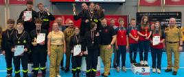 Uczestnicy II Halowych Młodzieżowych Zawodów Sportowo-Pożarniczych pozują na podium z medalami, pucharami i dyplomami po zakończonej rywalizacji.