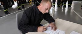 Strażak siedzi przy stoliku i podpisuje dokument. W tle widać innych strażaków oraz samochody pożarnicze