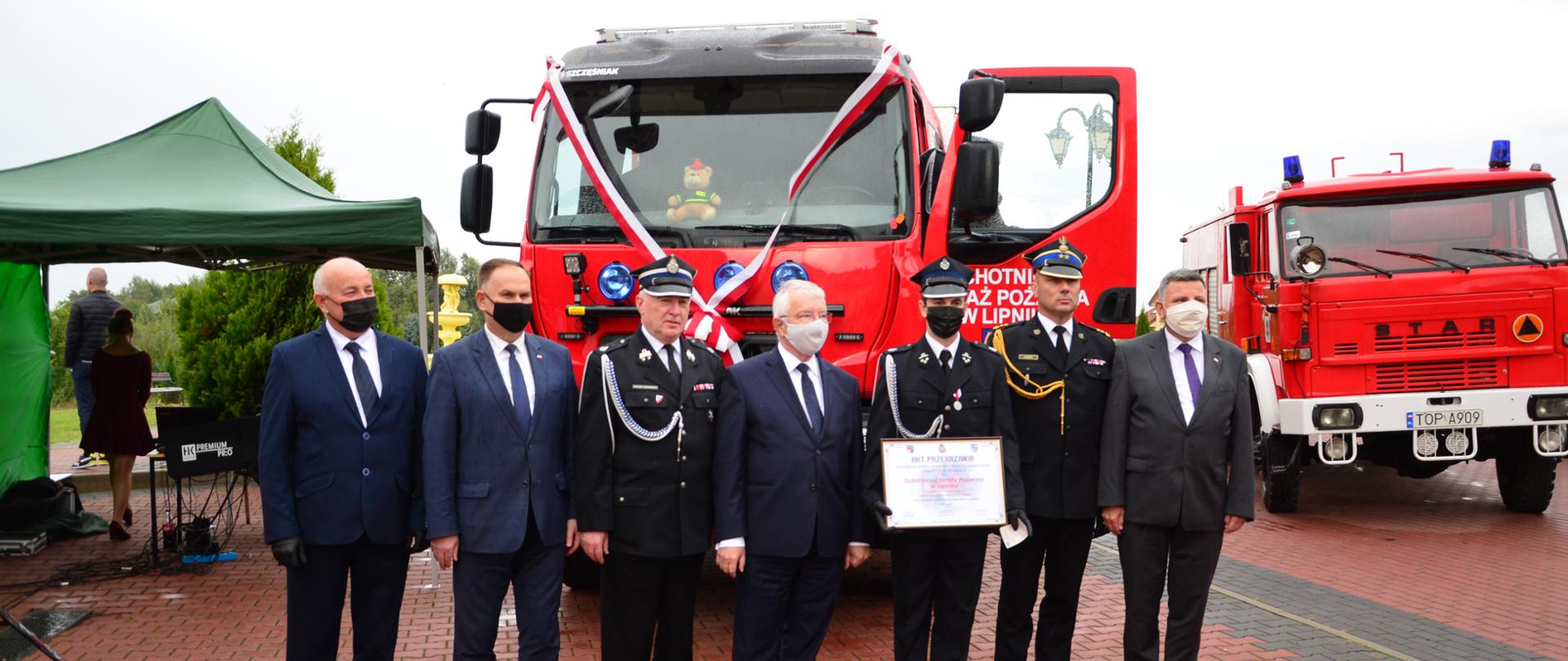 Czterech mężczyzn w garniturach i trzech strażaków w mundurach wyjściowych i rogatywkach stojący w szeregu przed nowo przekazanym samochodem gaśniczym. Jeden ze strażaków trzyma przed sobą akt przekazania pojazdu.