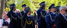 Grupa funkcjonariuszy w galowych mundurach straży pożarnej i policji salutuje podczas uroczystości na świeżym powietrzu. W tle widoczni są uczestnicy wydarzenia, w tym osoby w strojach ludowych oraz drzewa w słoneczny dzień.