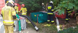 Zdjęcie przedstawia strażaków podczas ćwiczeń. Na zdjęciu widać zbiorniki na wodę oraz węże strażackie i samochód strażacki. 