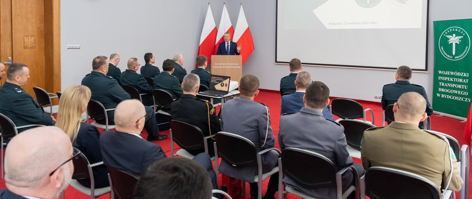 Jedna osoba stojąca przy mównicy i wypowiadająca się. Przed nim wiele siedzących wiele osób