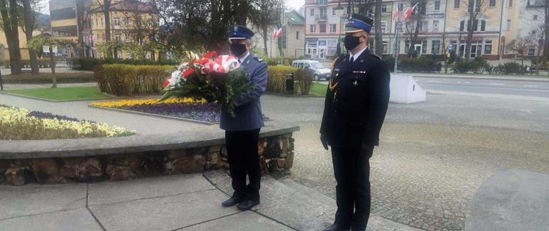 Komendant powiatowy państwowej straży pożarnej Emil Szmyd wraz z komendantem powiatowym policji Aleksandrem Szymaszkiem składają wiązankę pod Pomnikiem Żołnierza Polskiego w tle rynek w Ustrzykach Dolnych oraz zabudowa miasta.