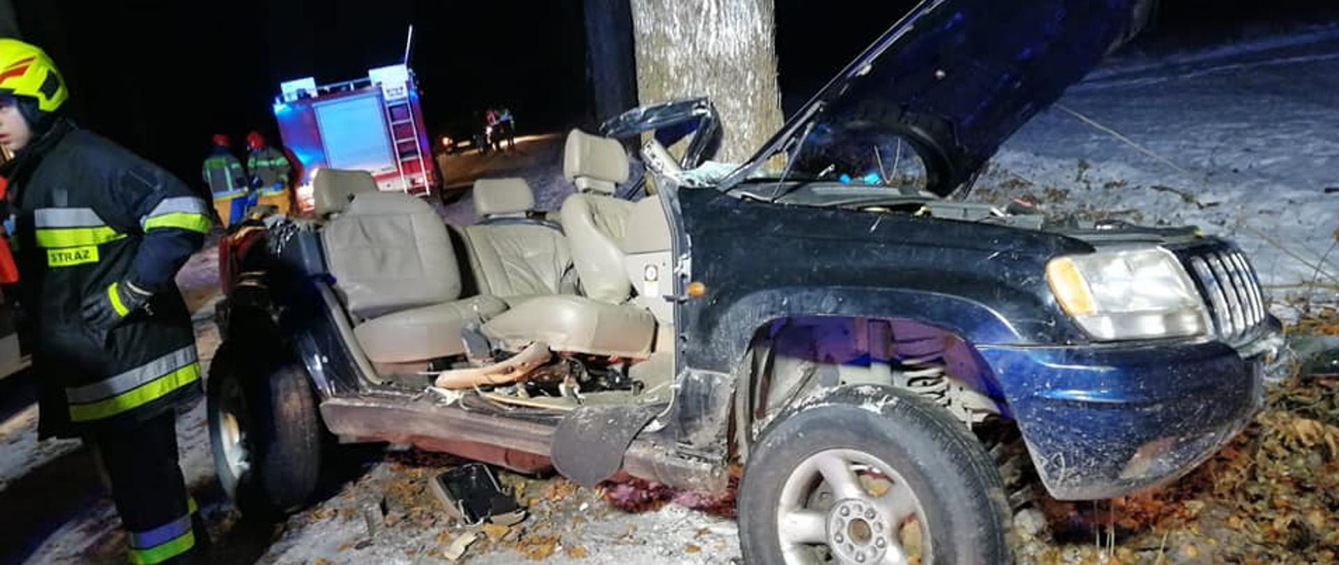 Zdjęcie przedstawia niebieski rozbity samochód JEEP, który ma odcięty dach, wnętrze pojazdu ma jasny kolor. Znajduję się on przy drzewie. Dookoła widać śnieg. Obok auta stoi strażak w żółtym hełmie oraz ciemnym ubraniu specjalnym. Za rozbitym samochodem znajduje się wóz strażacki. Zdjęcie zrobiono w porze nocnej. 