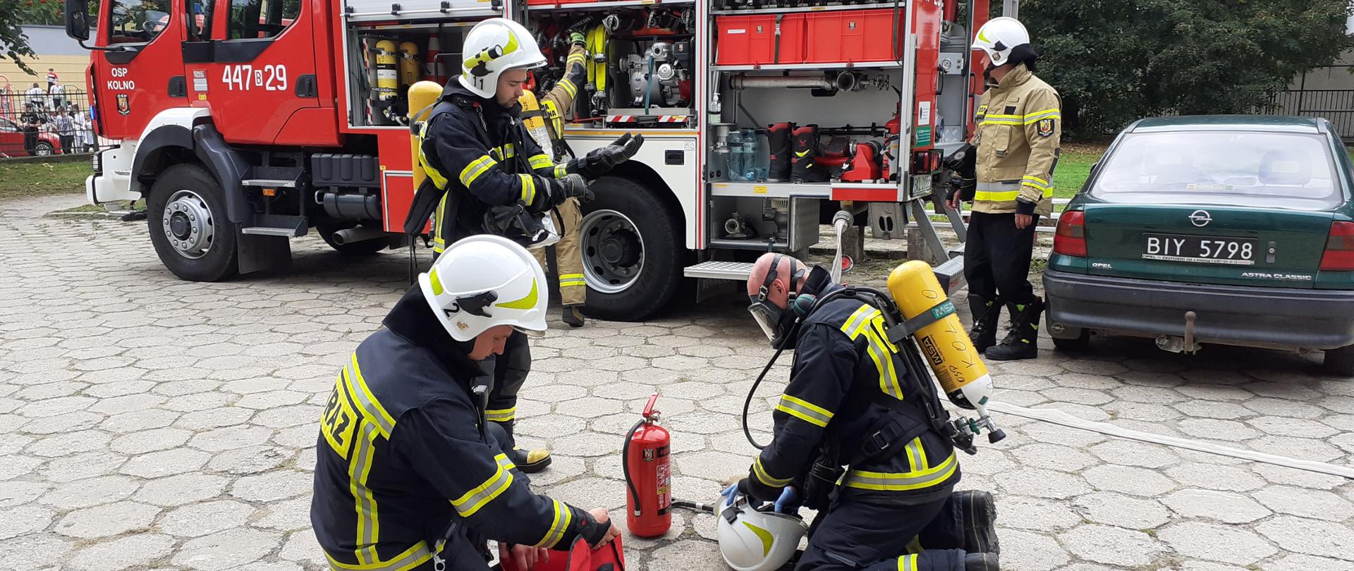 Widać strażaków przygotowujących sprzęt, w tle widać pojazd pożarniczy.