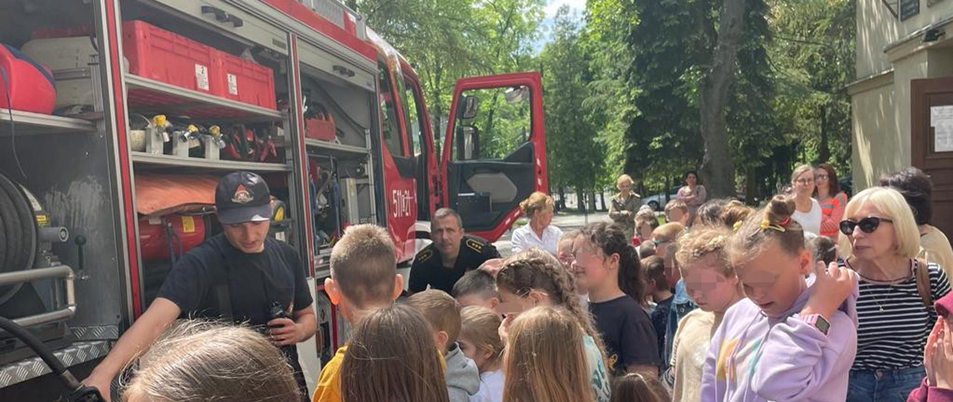 Zdjęcie wykonane jest przy samochodzie strażackim, na którym widać strażaków omawiających sprzęt pożarniczy dzieciom. Pojazd ma otwarte skrytki i widać w nim sprzęt pożarniczy. Jeden ze strażaków trzyma w ręku linię szybkiego natarcia. W tle zdjęcia widać park, oraz budynek szkoły, na którym wisi flaga Polski. 