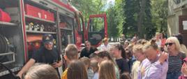 Zdjęcie wykonane jest przy samochodzie strażackim, na którym widać strażaków omawiających sprzęt pożarniczy dzieciom. Pojazd ma otwarte skrytki i widać w nim sprzęt pożarniczy. Jeden ze strażaków trzyma w ręku linię szybkiego natarcia. W tle zdjęcia widać park, oraz budynek szkoły, na którym wisi flaga Polski. 