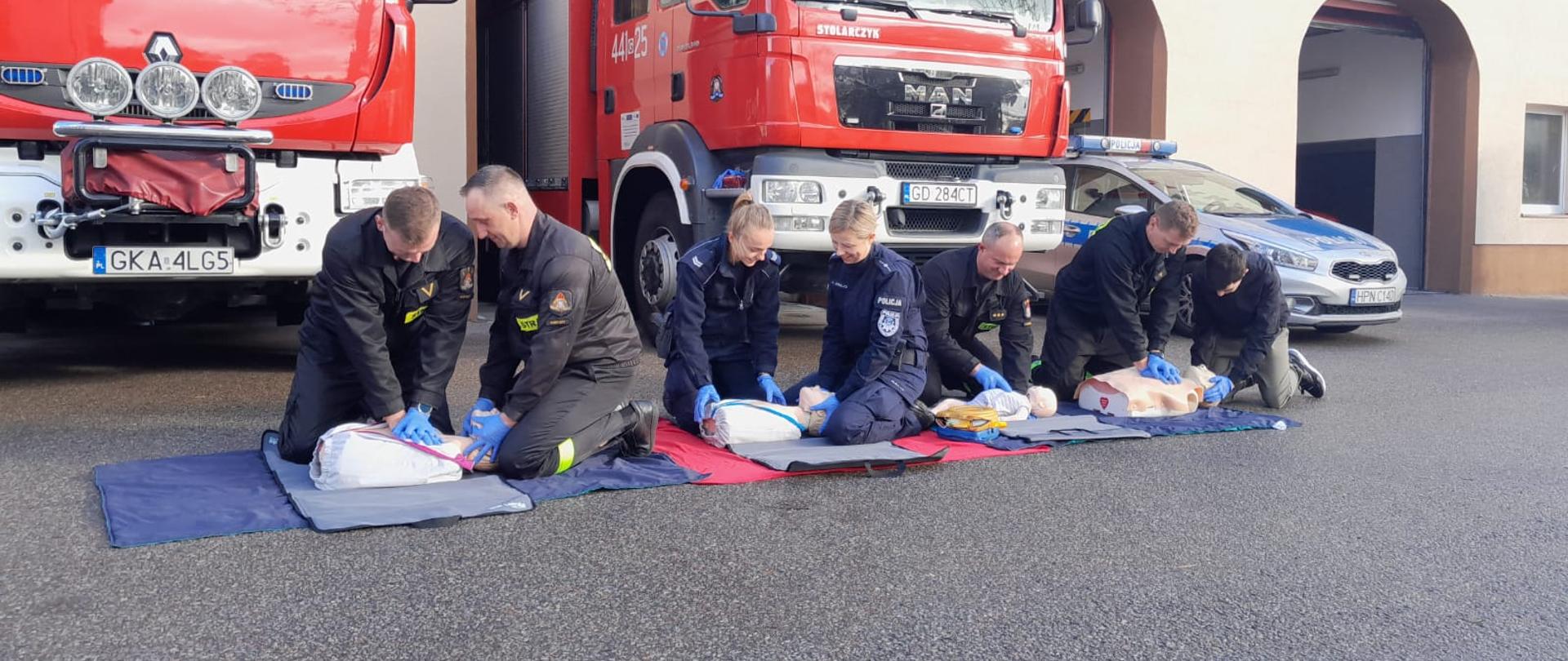 Zdjęcie przedstawia cztery stanowiska z fantomami, na których strażacy wspólnie z policjantkami prowadzą resuscytację. W tle pojazdy strażackie i radiowóz.