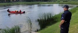 Zdjęcie przedstawia zbiornik wodny, na którym łodzią pływają strażacy wraz z policjantem zabezpieczeni w kamizelki asekuracyjne oraz kaski. Na brzegu stoi policjant, który obserwują całą akcję.