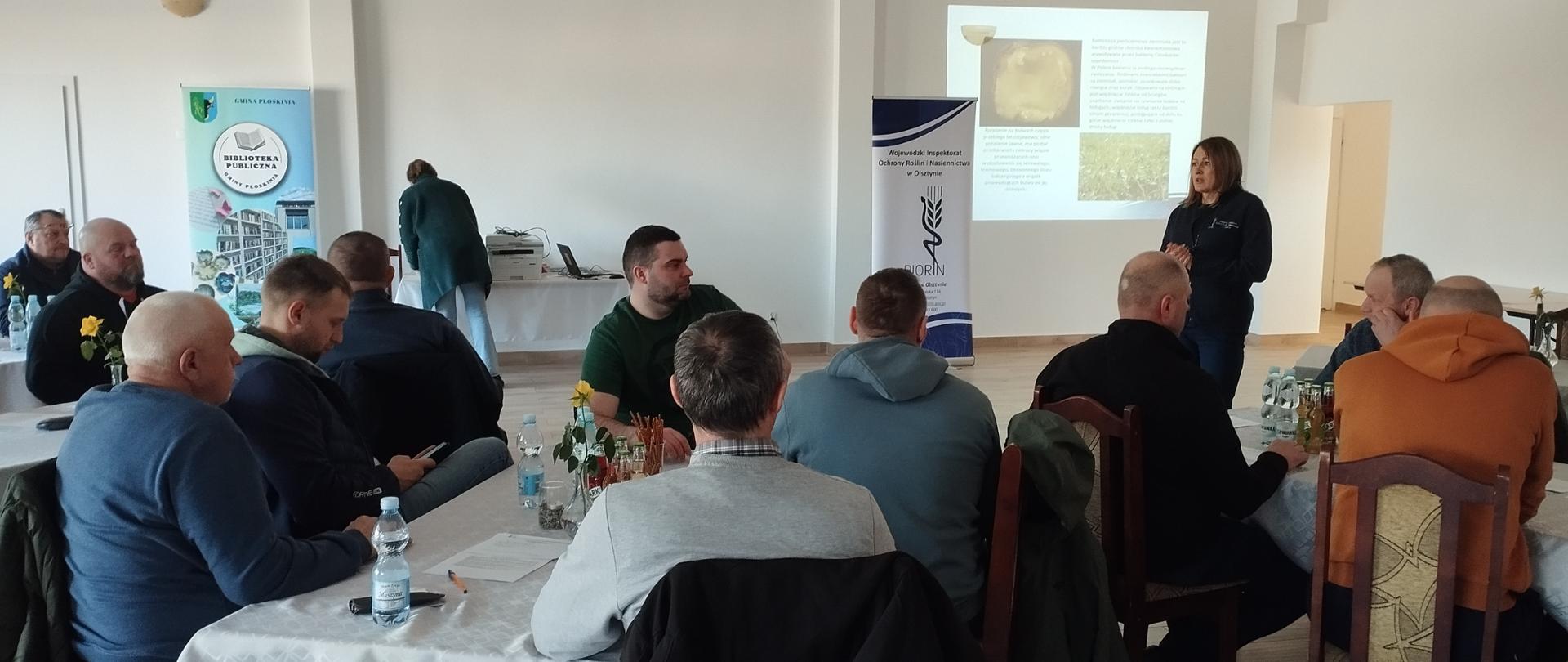 Kobieta stoi przed siedzącymi przy stolikach osobami, pośrodku jest baner WIORiN i slajd prezentacji