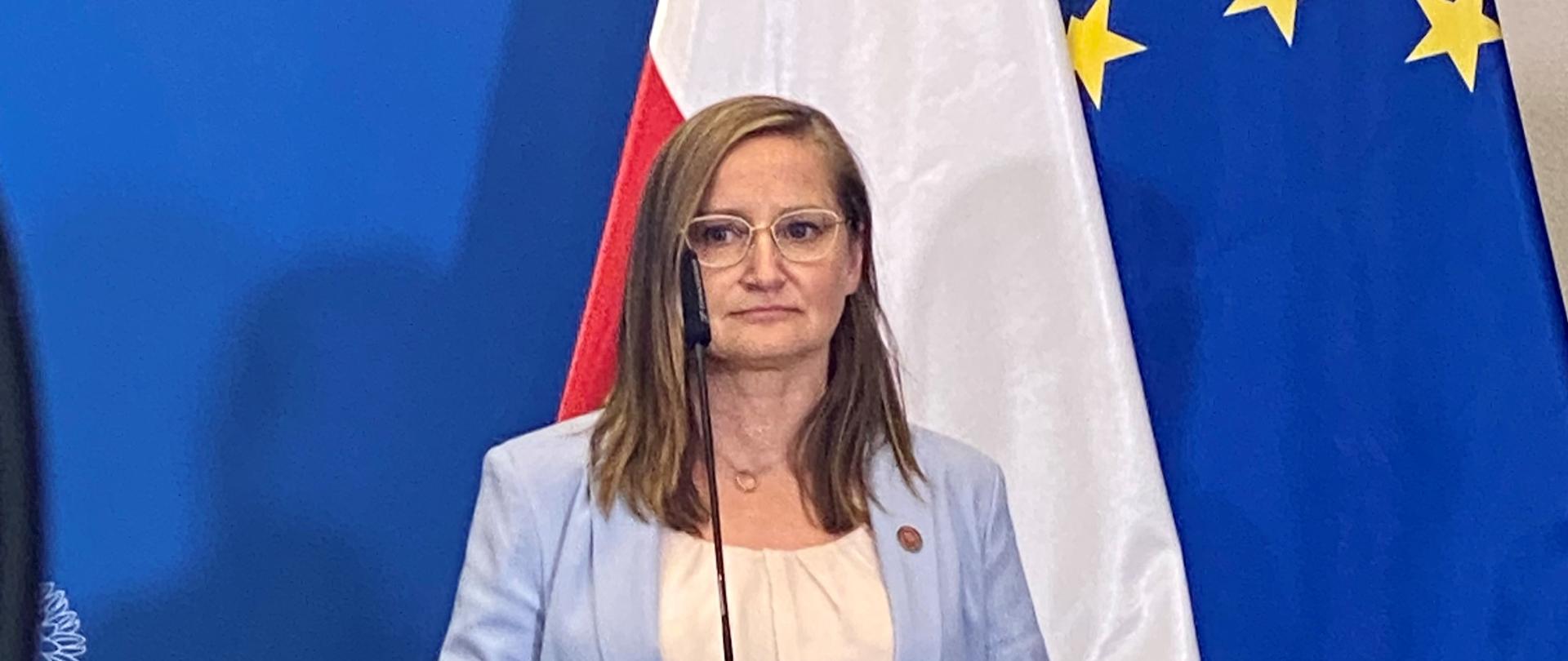 Kobieta stojąca przy mównicy w tle flagi polski i UE