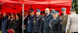 Fotografia przedstawia grupę oficjalnych gości oraz przedstawicieli służb mundurowych stojących pod zadaszeniem podczas uroczystego apelu z okazji Dnia Strażaka.