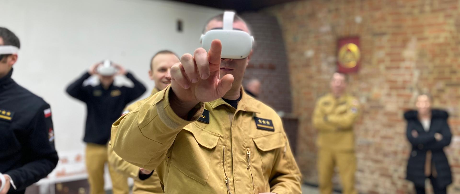 Na pierwszym planie strażak w mundurze służbowym z goglami VR na głowie/ Palcem wskazującym celuje w obiektyw. Za nim kilka osób ćwiczących. 