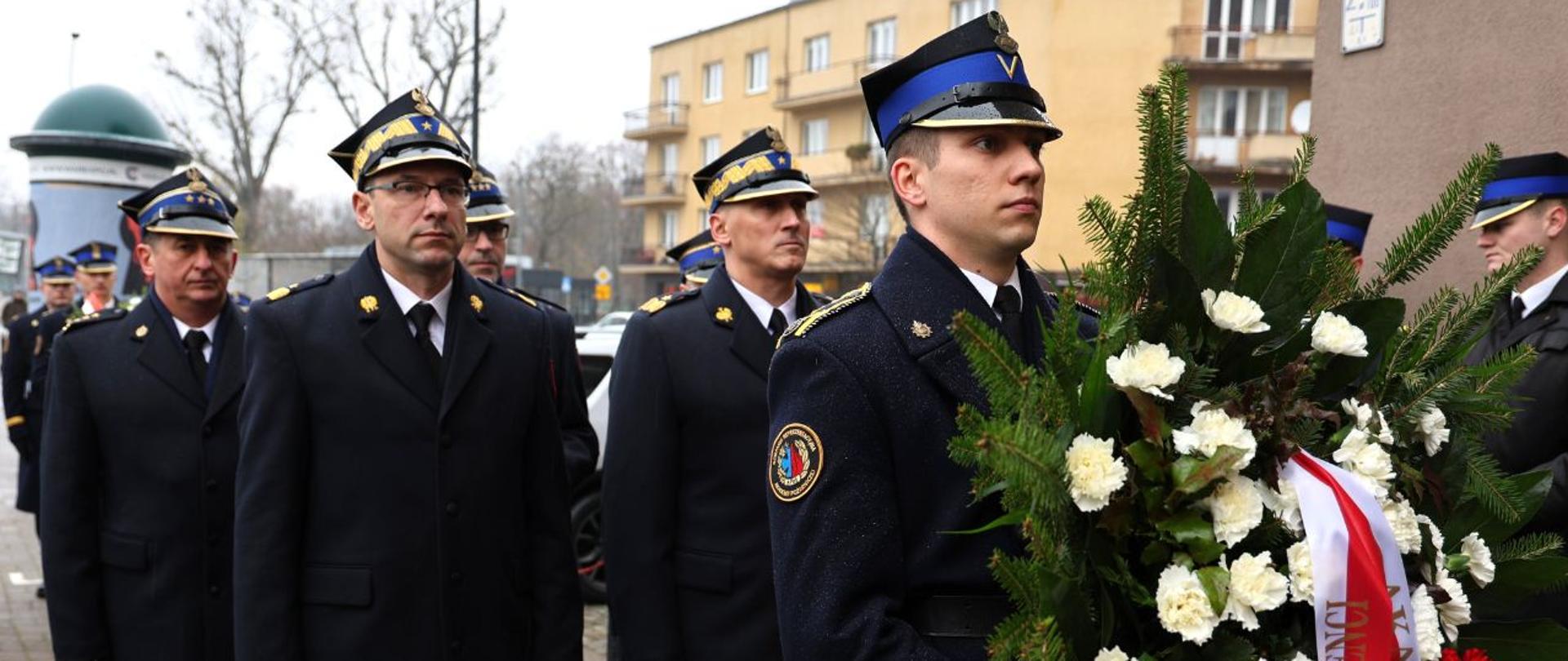 Funkcjonariusze w galowych mundurach stoją w szeregu, jeden z nich trzyma okazały wieniec z biało czerwonymi kwiatami.