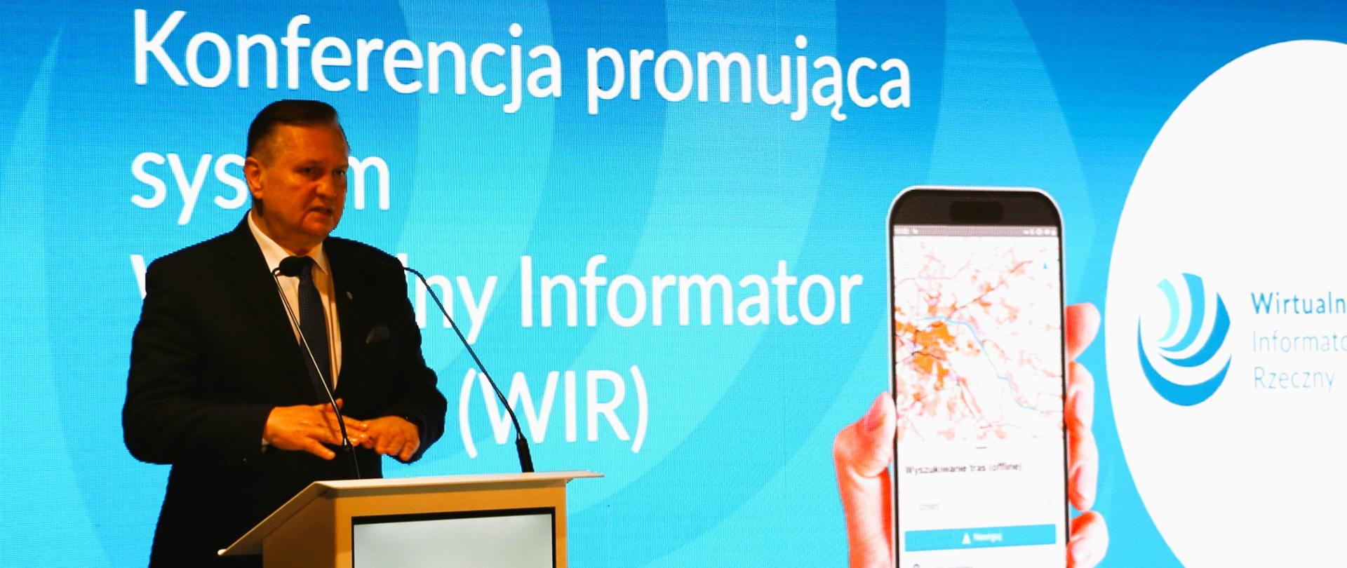 Prezes Wód Polskich - Krzysztof Woś stoi za mównicą i opowiada o Wirtualnym Informatorze Rzecznym. W tle na pierwszym planie dłoń trzymająca telefon komórkowy z widniejącą mapą żeglugową oraz slajd z napisem: Konferencja promująca system Wirtualny Informator Rzeczny. 