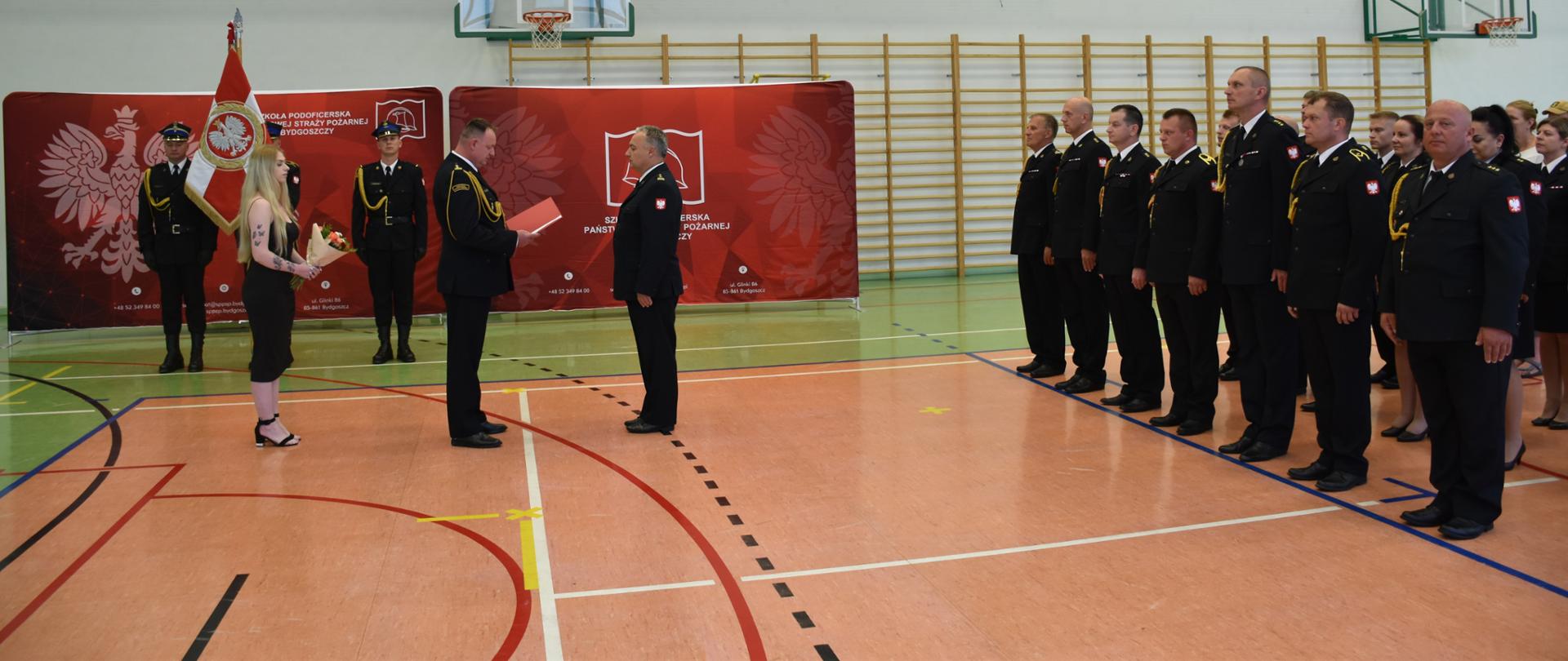Pamiątkowe zdjęcie grupowe z uroczystej zbiórki w Szkole Podoficerskiej PSP w Bydgoszczy z okazji powierzenia obowiązków Naczelnika Wydziału Nauczania bryg. Dariuszowi Jabłońskiemu
. Kadra i pracownicy szkoły pozują w hali sportowej na tle czerwonej ścianki SP PSP.