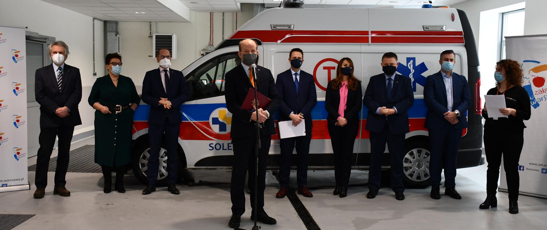 Elegancko ubrane kobiety oraz mężczyźni stoją na tle karetki pogotowia w pomieszczeniu szpitala