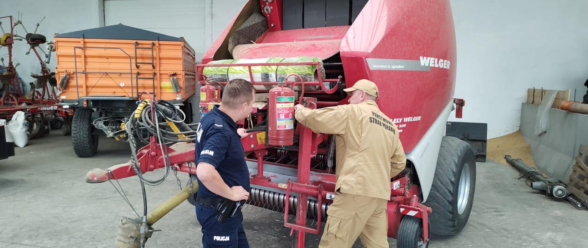 Przy czerwonej maszynie rolniczej - presie, stoi policjant i strażak, który sprawdza znajdującą się na maszynie gaśnicę.