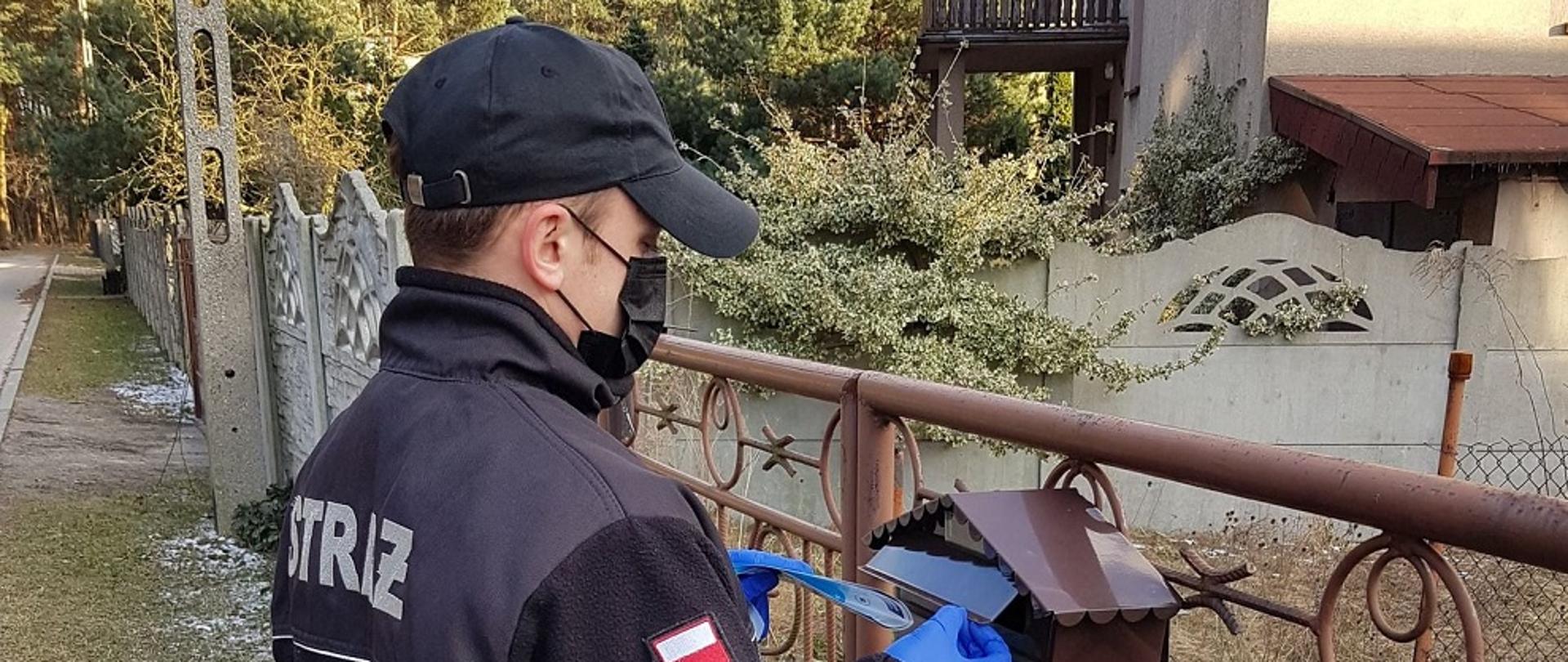 Zdjęcie przedstawia druha OSP w ubraniu koszarowym stojącego przed bramą prywatnej posesji podczas kolportażu ulotek informacyjnych na temat profilaktyki oraz szczepień przeciwko COVID-19