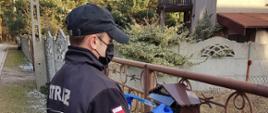 Zdjęcie przedstawia druha OSP w ubraniu koszarowym stojącego przed bramą prywatnej posesji podczas kolportażu ulotek informacyjnych na temat profilaktyki oraz szczepień przeciwko COVID-19