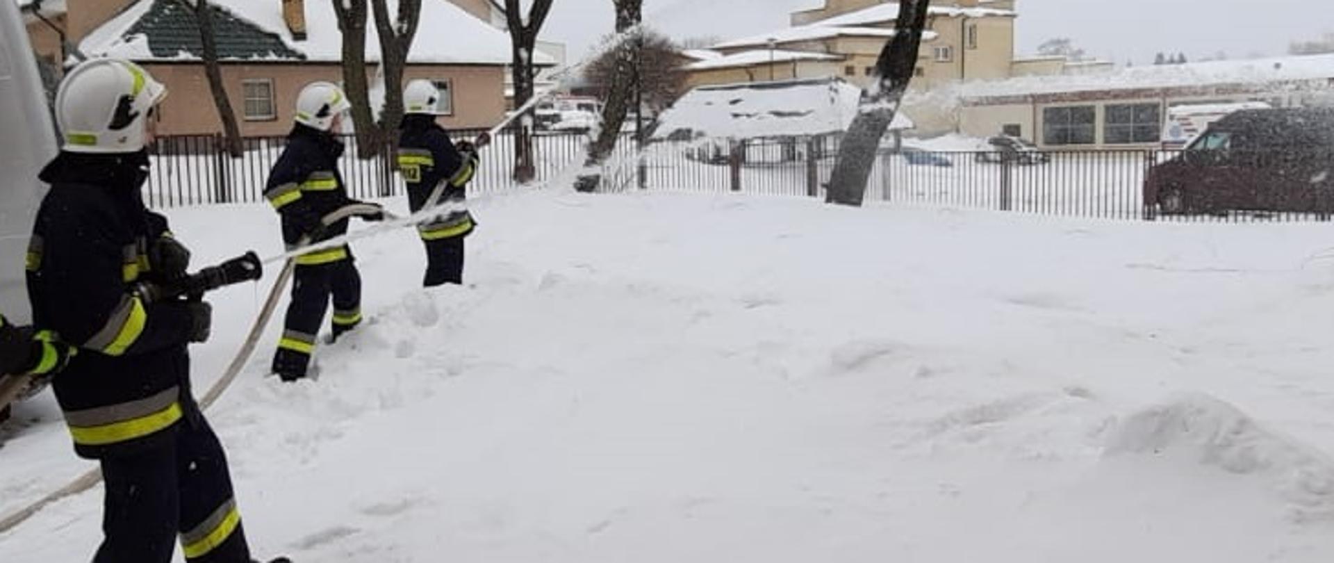 Zdjęcie obrazuje 3 strażaków trzymających węże z których leci woda na ziemi leży śnieg w tle widać drzewa i budynki