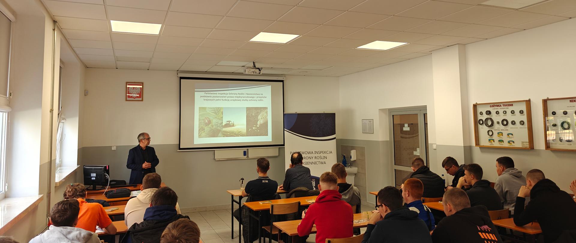 Uczniowie w sali lekcyjnej siedzą w ławkach. Słuchają prezentacji pracownika WIORiN Pana Andrzeja Borodziuka.