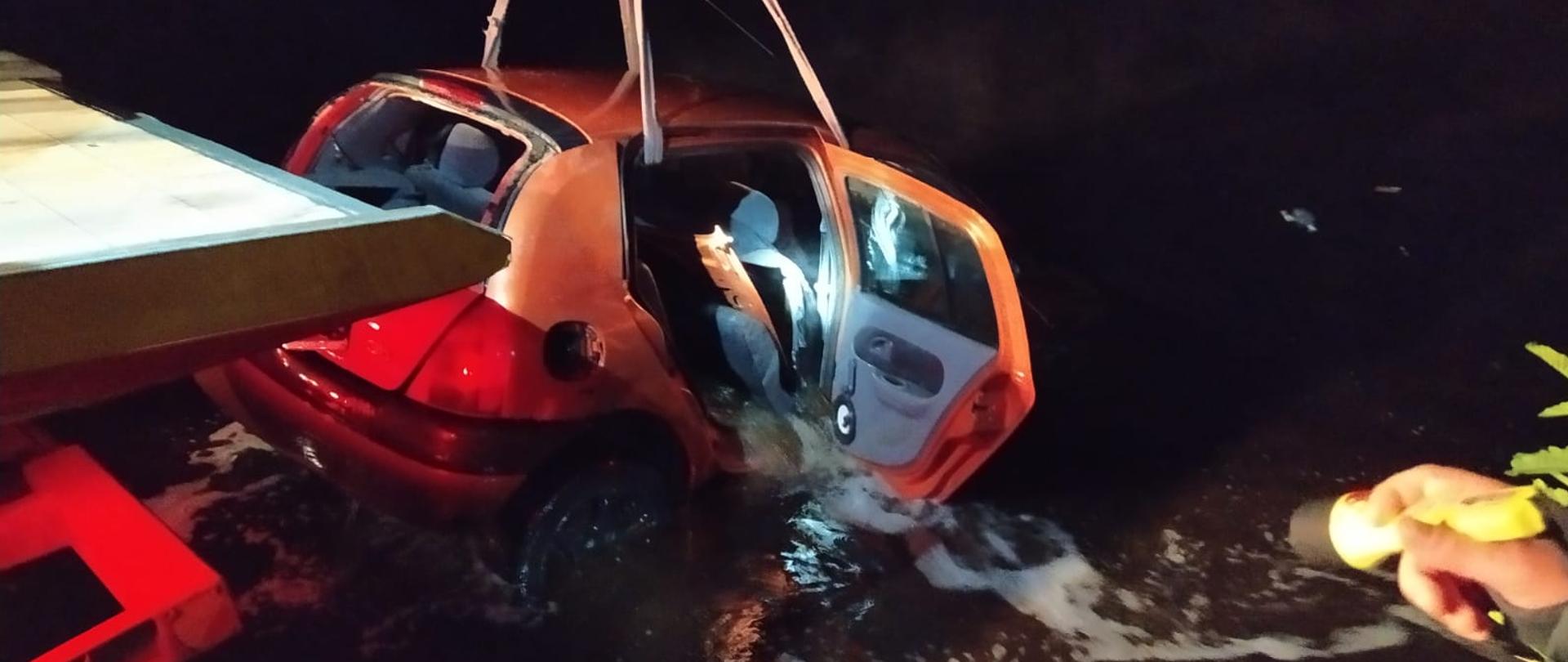 Zdjęcie przedstawia czerwony samochód wyciągany z jeziora przy pomocy dźwigu.