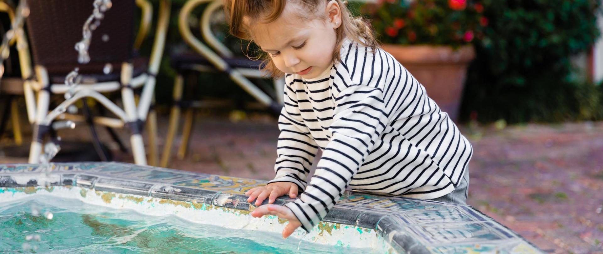 Na zdjęciu widać dziewczynkę zanurzającą swoją rękę w wodzie z fontanny.