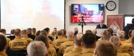 Mężczyzna stoi przy mównicy i przemawia do funkcjonariuszy którzy siedzą na krzesłach na ekranach wyświetlana jest prezentacja.