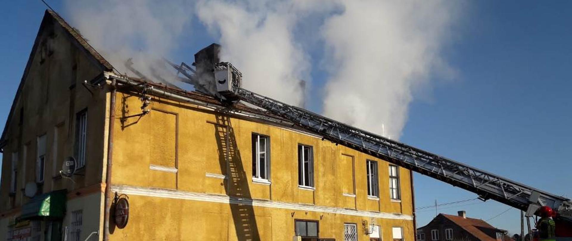 Widoczny jest dach budynku wielorodzinnego w Słoneczniku. Pomimo ugaszenia płomieni nadal wydobywają się z dachu duże ilości dymu. Trwa dogadzanie dachu przy pomocy wysięgnika samochodu ratowniczo-gaśniczego.