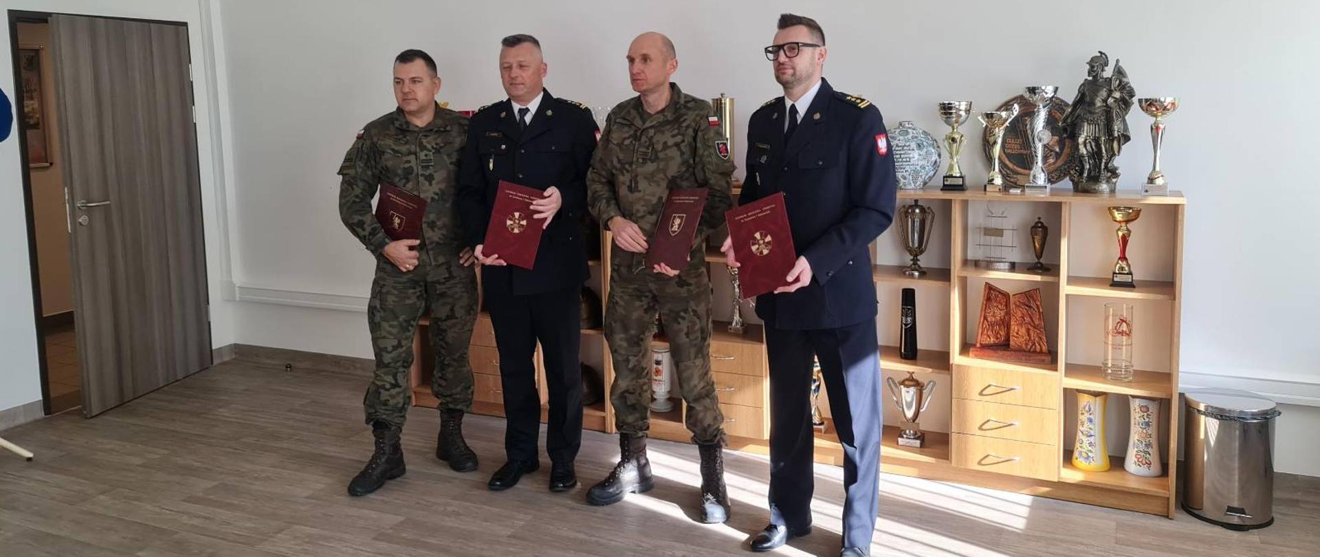 Zdjęcie przedstawia 4 mężczyzn w mundurach stojących w szeregu przed gablotą z pucharami. Dwóch z nich ubranych jest w mundury moro, dwaj pozostali w czarne galowe mundury. W tle ściana z napisem „Szkoła Specjalistów Pożarnictwa”