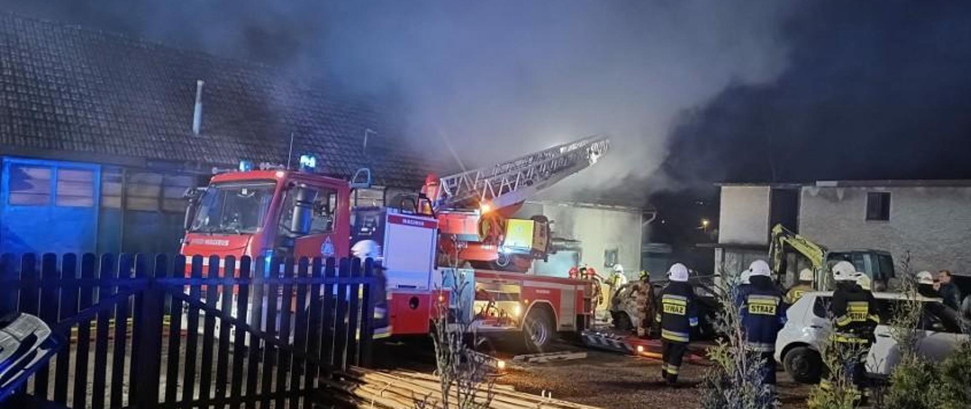 W środku kadru, znajduje się mechaniczna drabina pożarnicza. Samochód ma rozłożoną drabinę w stronę palącego się budynku gospodarczego. Wokół drabiny widać kłęby dymu, w tle widać budynek gospodarczy objęty pożarem. W około drabiny widać rozproszoną grupę strażaków.