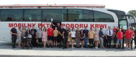 Na zdjęciu osoby biorące udział w akcji krwiodawstwa na tle białego autokaru, który służy za mobilny punkt poboru krwi.