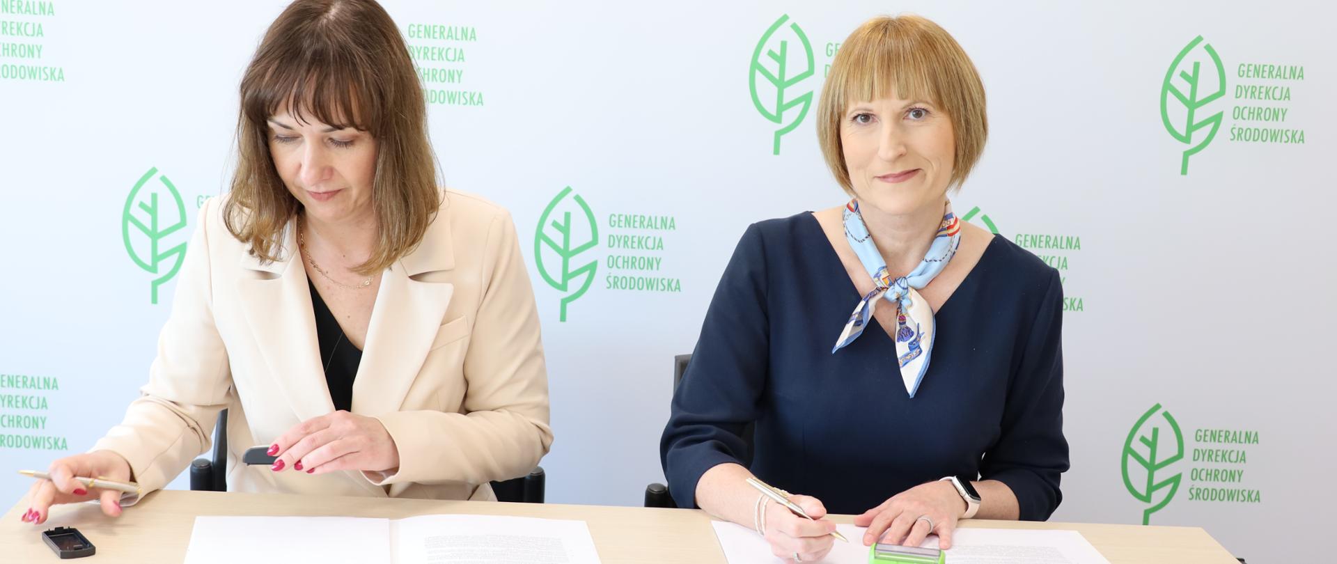 Na tle białej ściany z logotypem Generalnej Dyrekcji Ochrony Środowiska (zielony liść) siedzą dwie kobiety. Podpisują dokumenty. 
