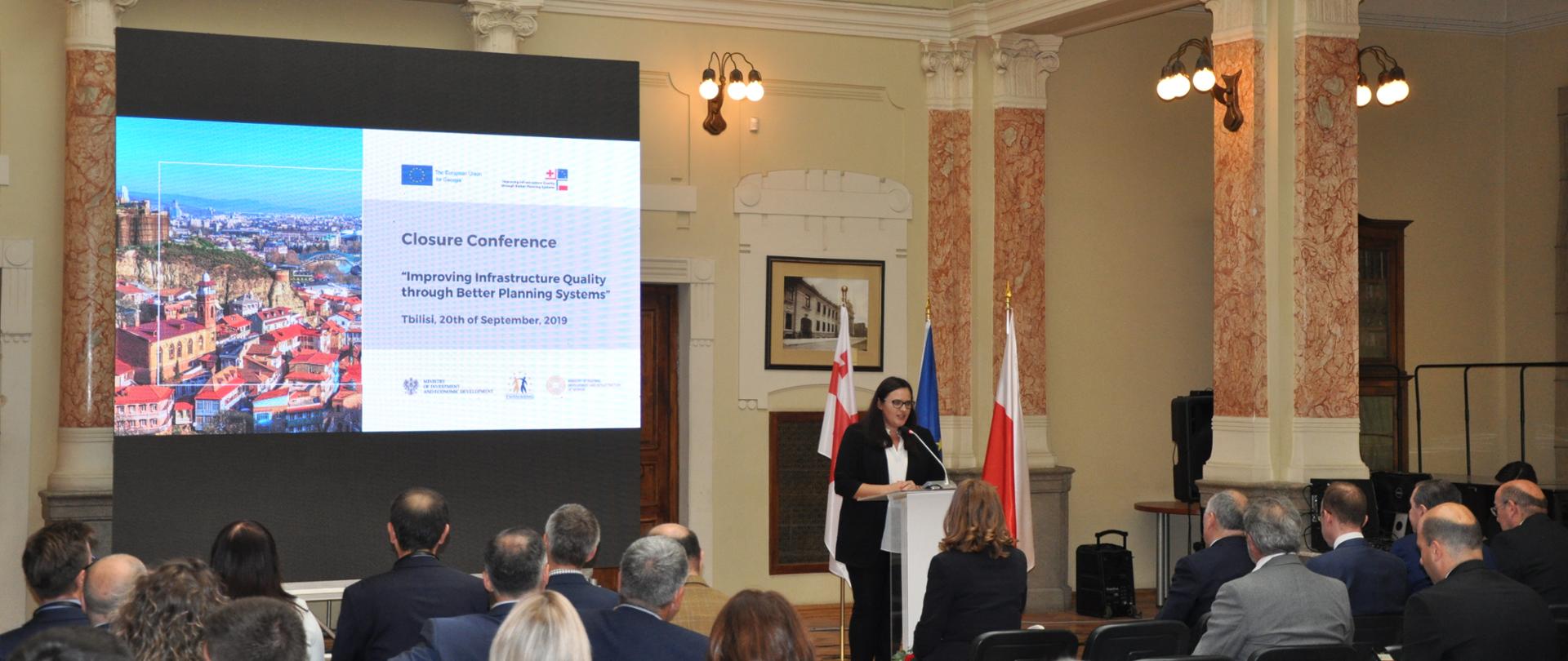 W sali siedzą uczestniczy konferencji. Przodem do nich w mównicy przemawia wiceminister Jarosińsk-Jedynak. Za jej plecami wyświetlona jest prezentacja.