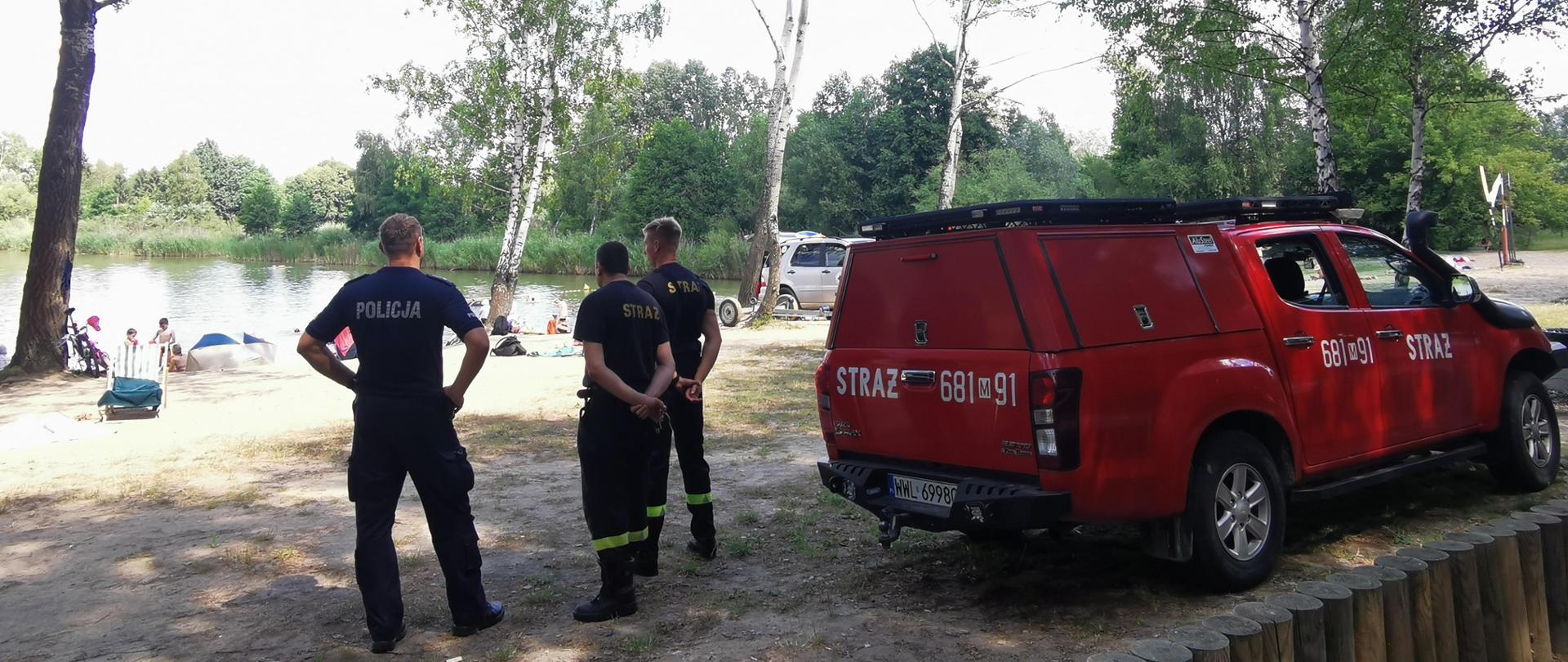 Zdjęcie przedstawia piaszczystą plażę na której rośnie kilka drzew. Na pierwszym planie widać policjanta i dwóch strażaków. Obok stoi czerwony samochód strażacki. Na drugim planie widać zażywających słonecznych kąpieli ludzi.
