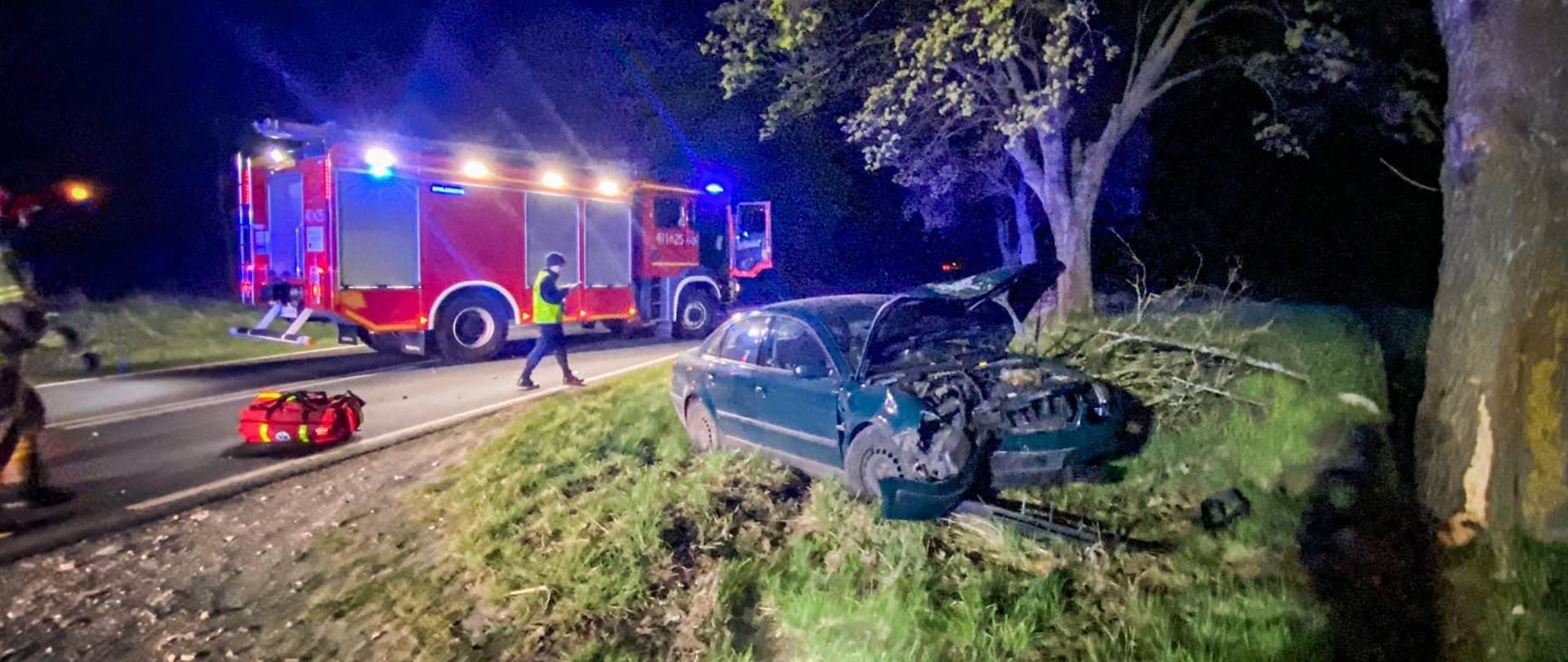 Na zdjęciu widać samochód osobowy, który uderzył w drzewo. W tle samochód straży pożarnej.
